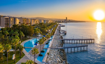 Promenada nadmorska w Limassol na Cyprze o zachodzie słońca; rząd palm, nowoczesne budynki wzdłuż wybrzeża, pomosty wychodzące w morze i spokojna tafla wody odbijająca słońce.