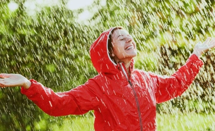dziewczyna w czerwonej przeciwdeszczowej kurtce cieszy się z deszczu w słońcu. Ma uniesione ręce.